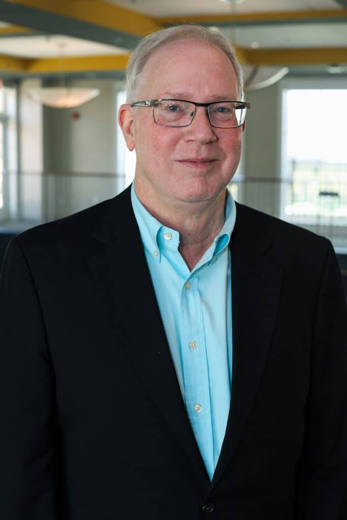 John McManus, wearing glasses and dressed in a black blazer and light blue shirt, standing inside a building.
